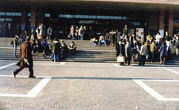 A train station in Venice in 1977. Crowds of people sit and stand on the stairs.