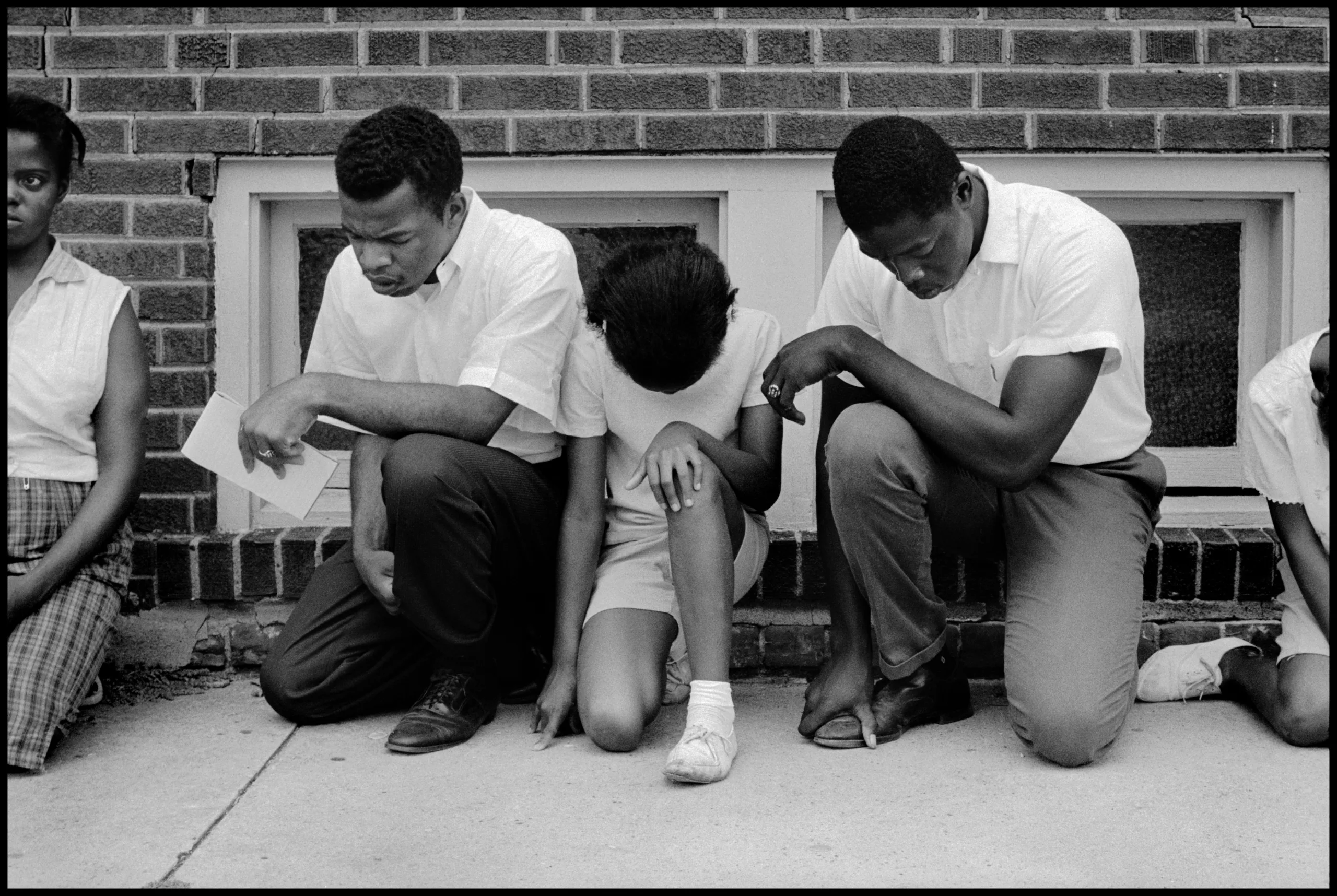 Black-and-white photo of three young African Americans one knee in prayer with their heads down in front of a brick building. On the left and right are two men who look to be in their early 20s. There is a girl in the middle.