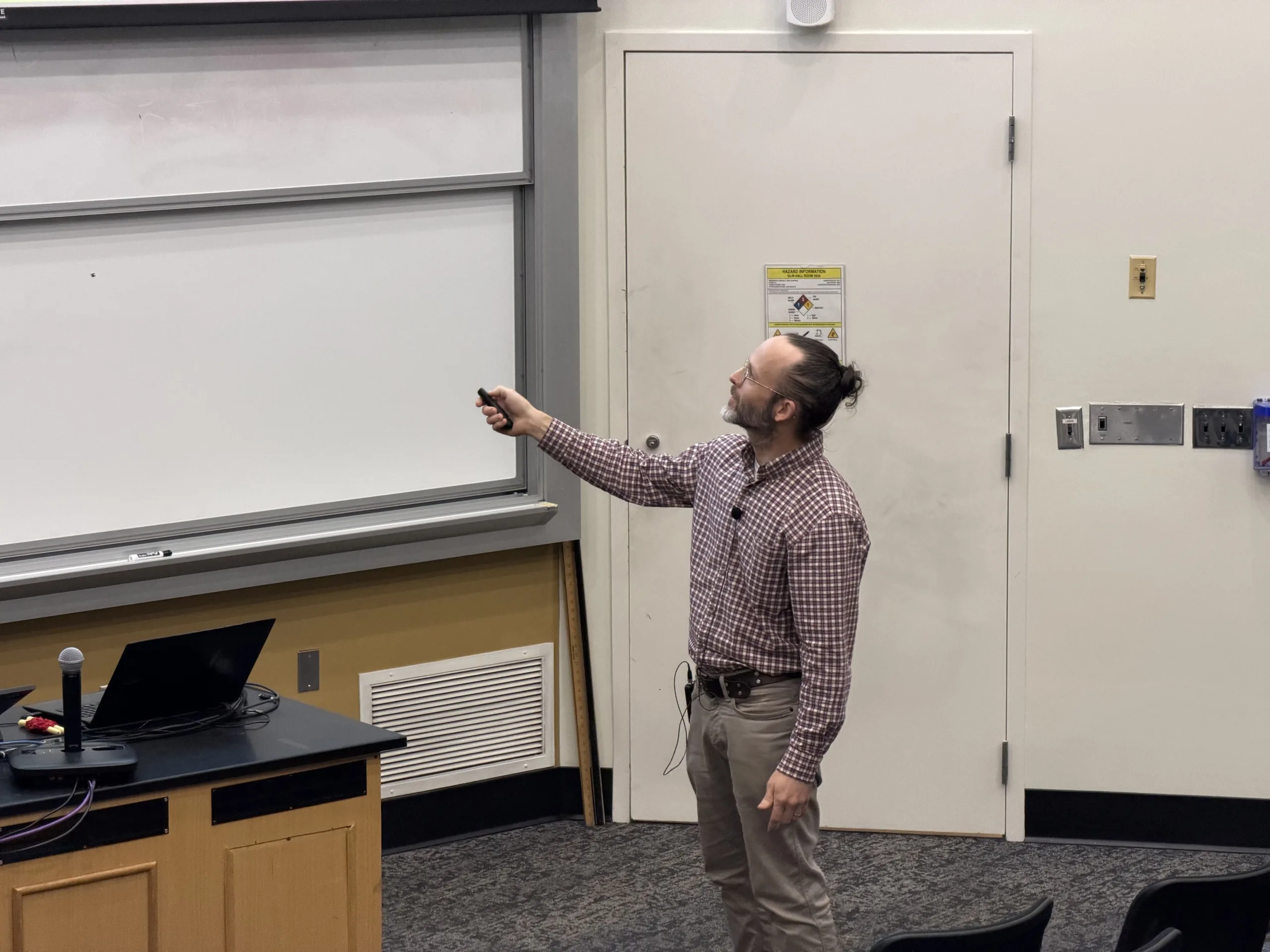 Lee Pryor gestures toward the projection screen with a remote at the front of a lecture room.