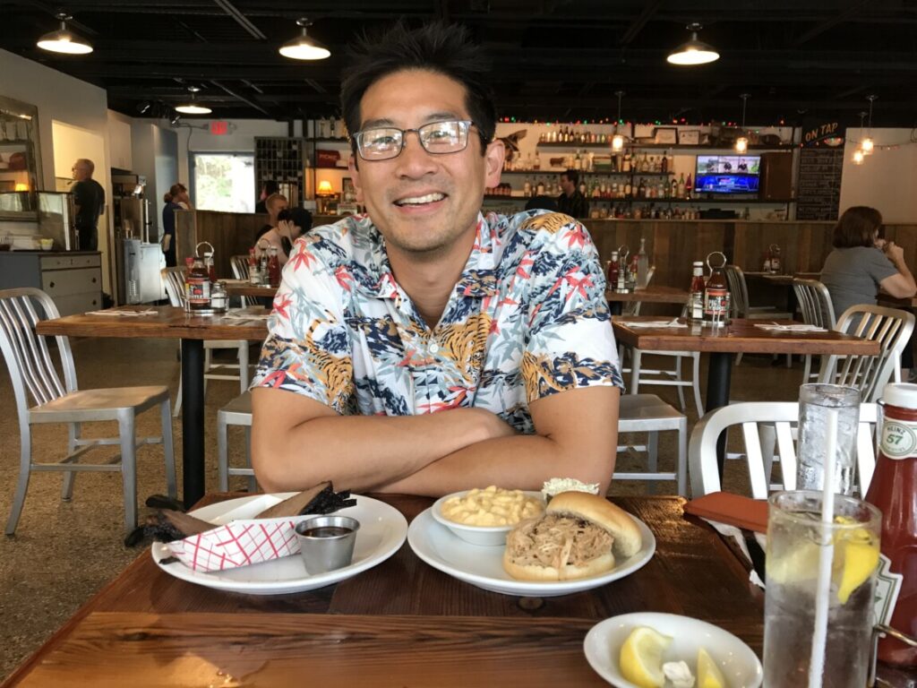 Professor Derek P. Lee sits a table with a barbecue sandwich in front of him. He's smiling and wearing a Hawaiian-print shirt. 