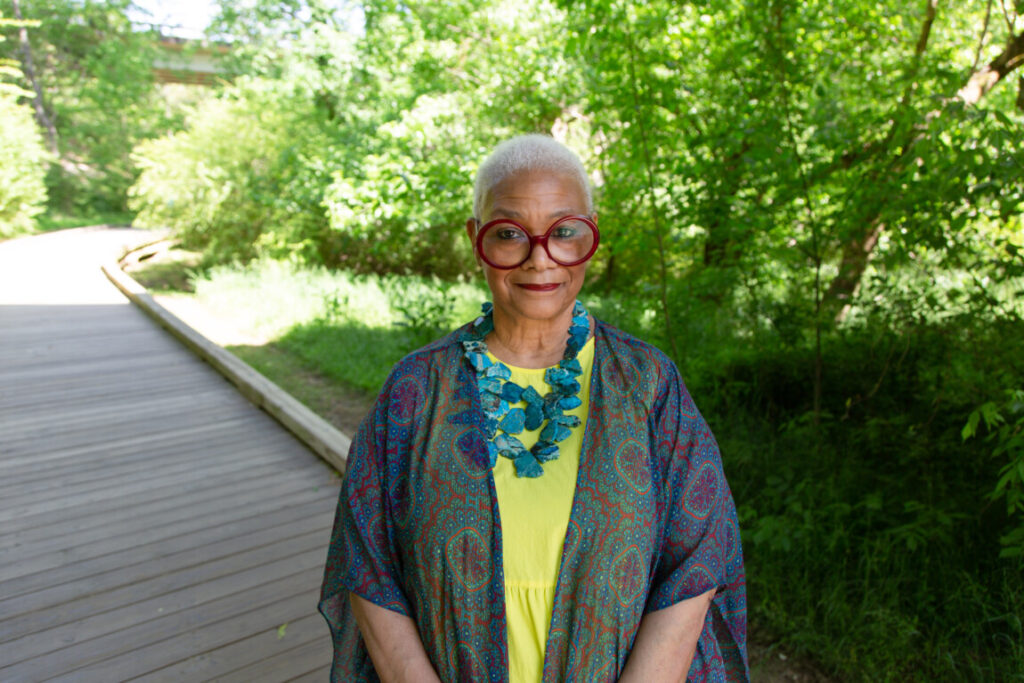 Poet Jaki Shelton Green stands on a wooden walkway with trees behind her.