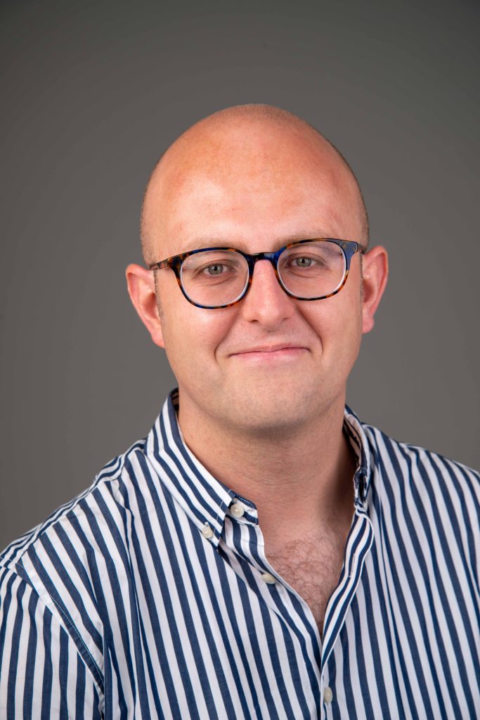 An image in color of Nathan Waddell from the chest up. He is a bald white man and is wearing a striped button-up shirt and glasses.
