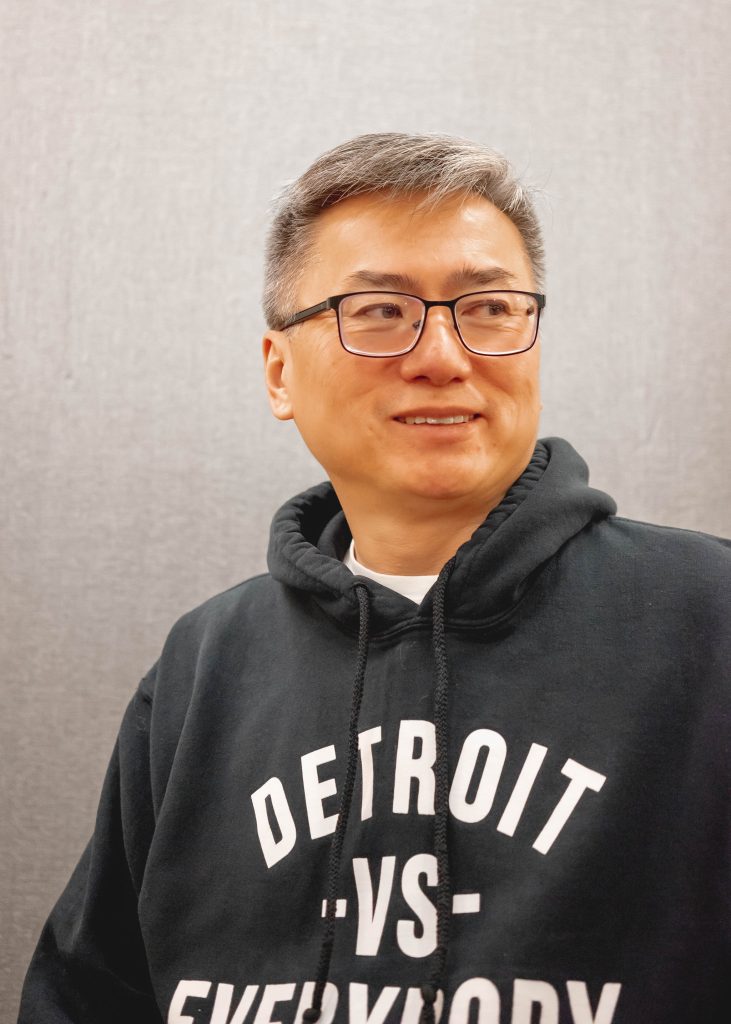 Curtis Chin appears from the chest up, wearing glasses and a hoodie with Detroit VS Everybody written on it. He looks to the side of the camera.