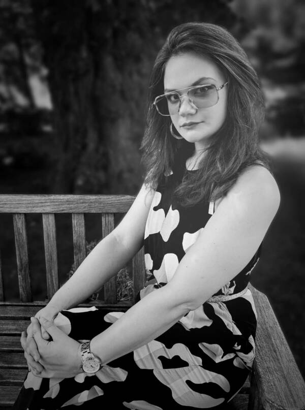 Holly, a young white woman wearing an abstract-patterned dress and glasses, is depicted in black and white.