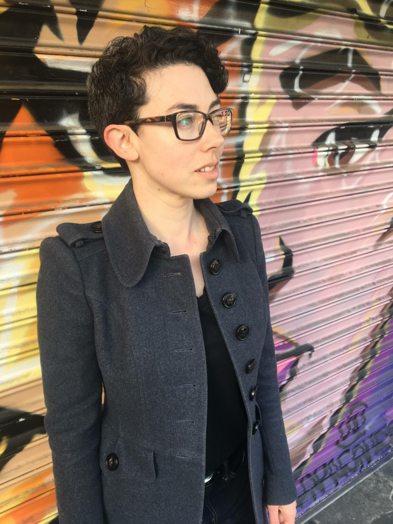 Dr. Lorden, a woman with short dark hair and rectangular glasses, stands in front of a wall. A painted mural on the wall depicts half of a face.