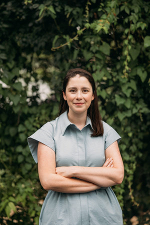 A photograph of Caroline Hallemann, a white woman wearing a blue dress and smiling at the camera. She stands with her arms crossed in front of a blurred background of green vines.