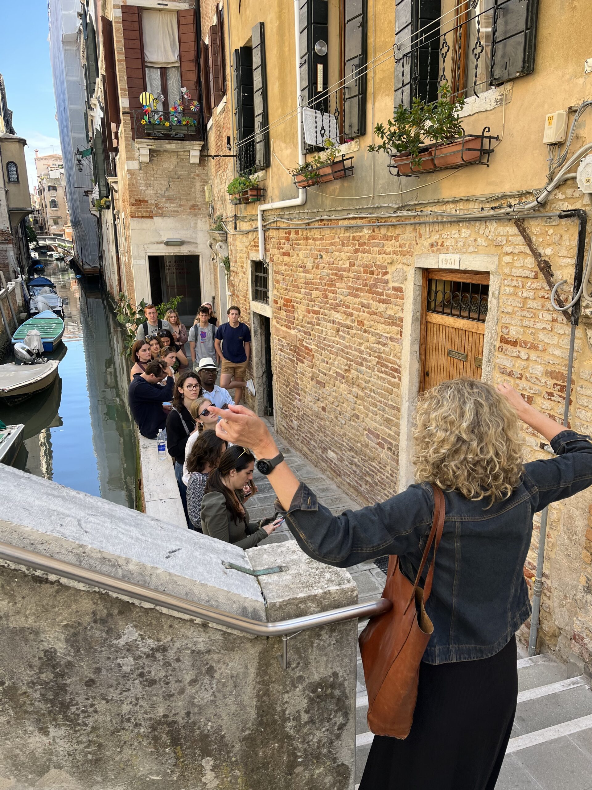 Venice, 2023: A professor with their back to the camera speaks to a group of students alongside a canal in Venice. 