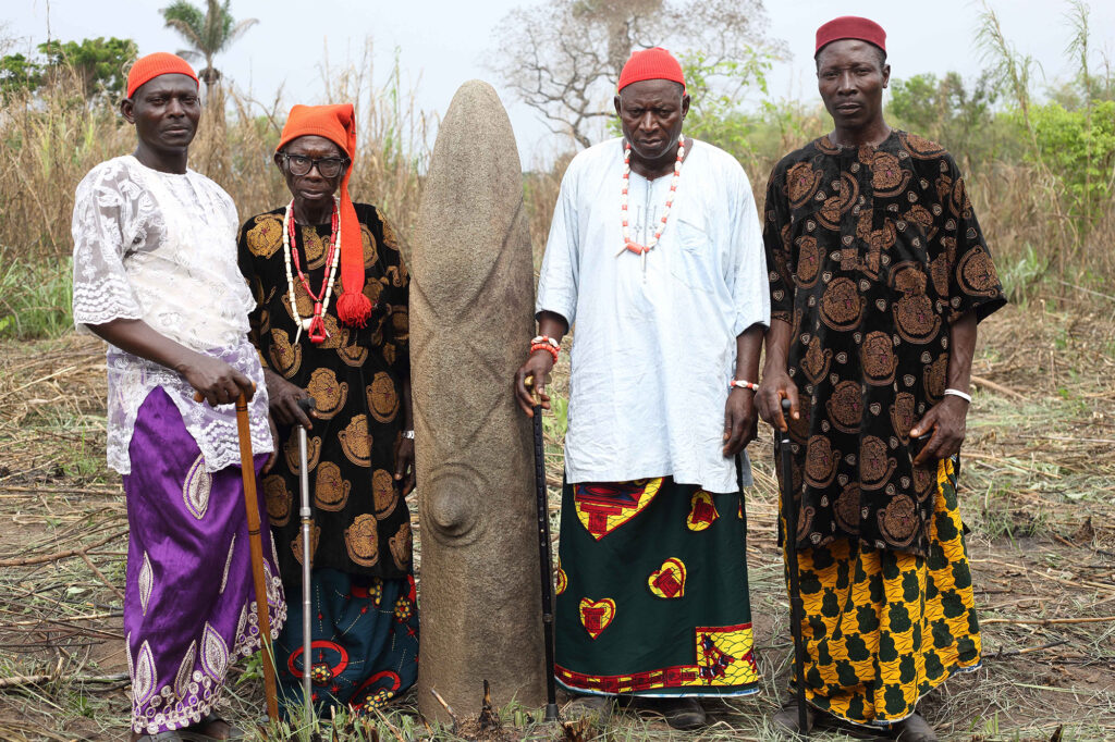 Four Chiefs of Oyengi stand with the last remaining carved Bakor monolith. The Bakor monoliths are venerated carved stone sculptures representative of ancestors, associated with traditional spiritual and social practices within the
forest belt of Cross River State, south-east Nigeria. They are locally known as “akwanshi” or “atal” and found as a collection of stone monoliths within family,clan, or village lands.  Photo credit: Ferdinand Saumarez Smith, 2018
