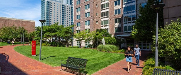 A photo of the Boston University housing option. An external shot of high-rise residential buildings, with a bright green, freshly mowed lawn in front, encircled by red brick walkways with black metal benches. Two people are walking along the walkway on the rigth.