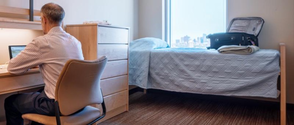 An internal photo of the Boston University housing option. A balding white man with graying black hair and a white checkered shirt sits in a tan chair at a light wooden desk, working at a laptop. To his right is a light wood chest of drawers. To the right of that, under a window with a view of the Boston skyline and a sky-blue sky, is a twin long bed with sky-blule bedcover. On top of the bed is an open black suitcase. The floor appears to have carpeted or laminate floors.