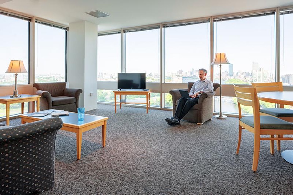 An interior photo of a lounge space at the Boston University housing option. An open carpeted space with various kinds of seating: easy chairs, coffee tables, a console table with a TV on it, an armchair with a side table and table lamp, a round table with chairs around it. Two walls have wall-to-wall, floor-to-ceiling windows overlooking the Boston skyline.