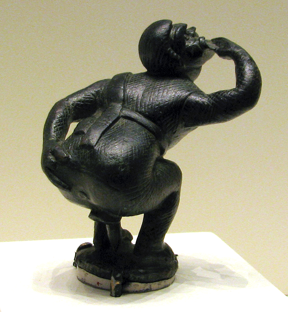 An ancient statuette of a comic actor. In black metal, the figure is bent at the knees and bent forward at the waist, head craned towards the sky, right hand on chin in a pensive gesture, left hand spreading open buttcheeks to reveal a prominent anus.