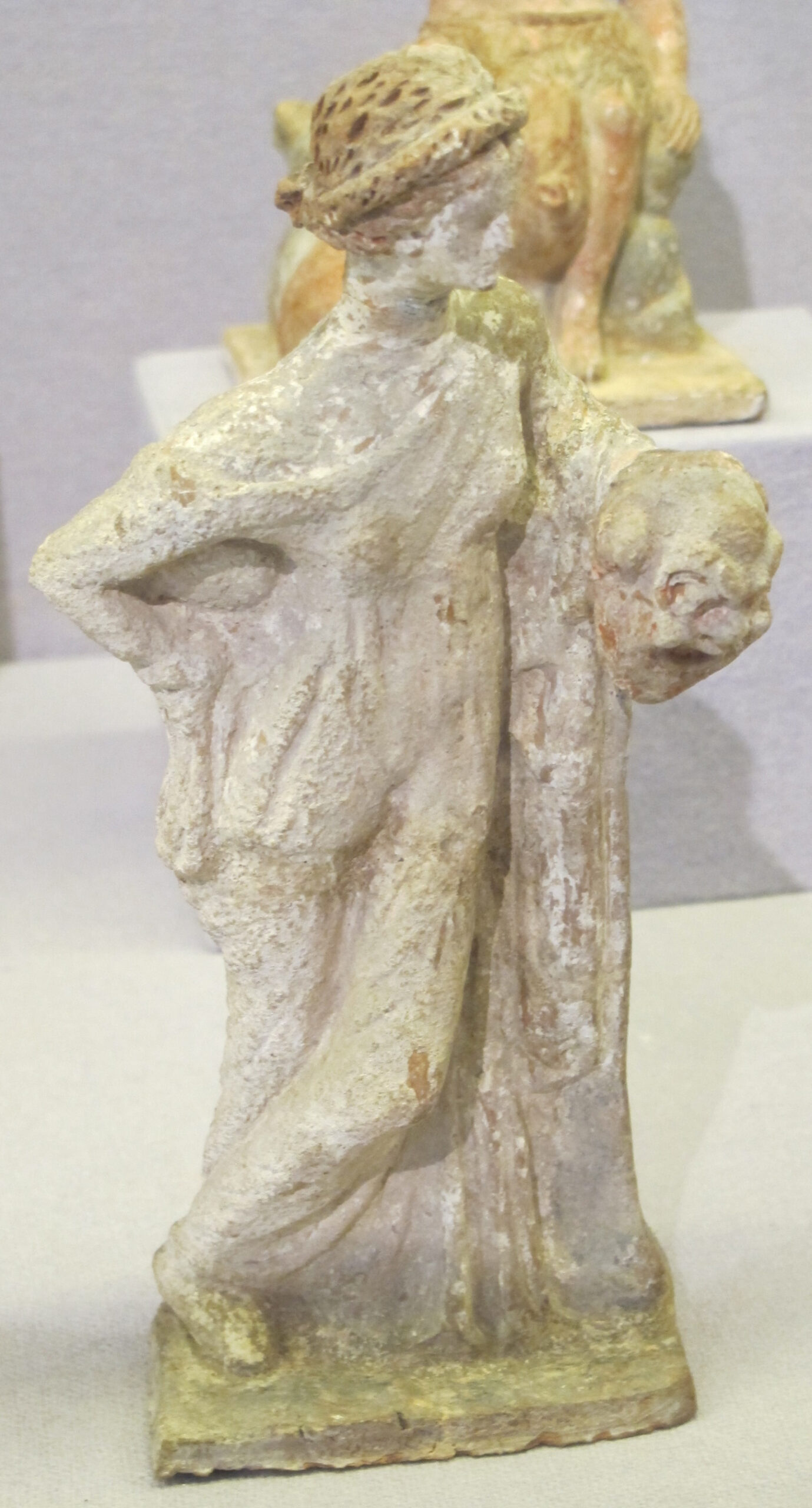 An ancient Greek terracotta figurine from Tanagra of the muse of comedy. A woman with brown braided hair leaning on her left side and holding a smiling mask in her left hand.