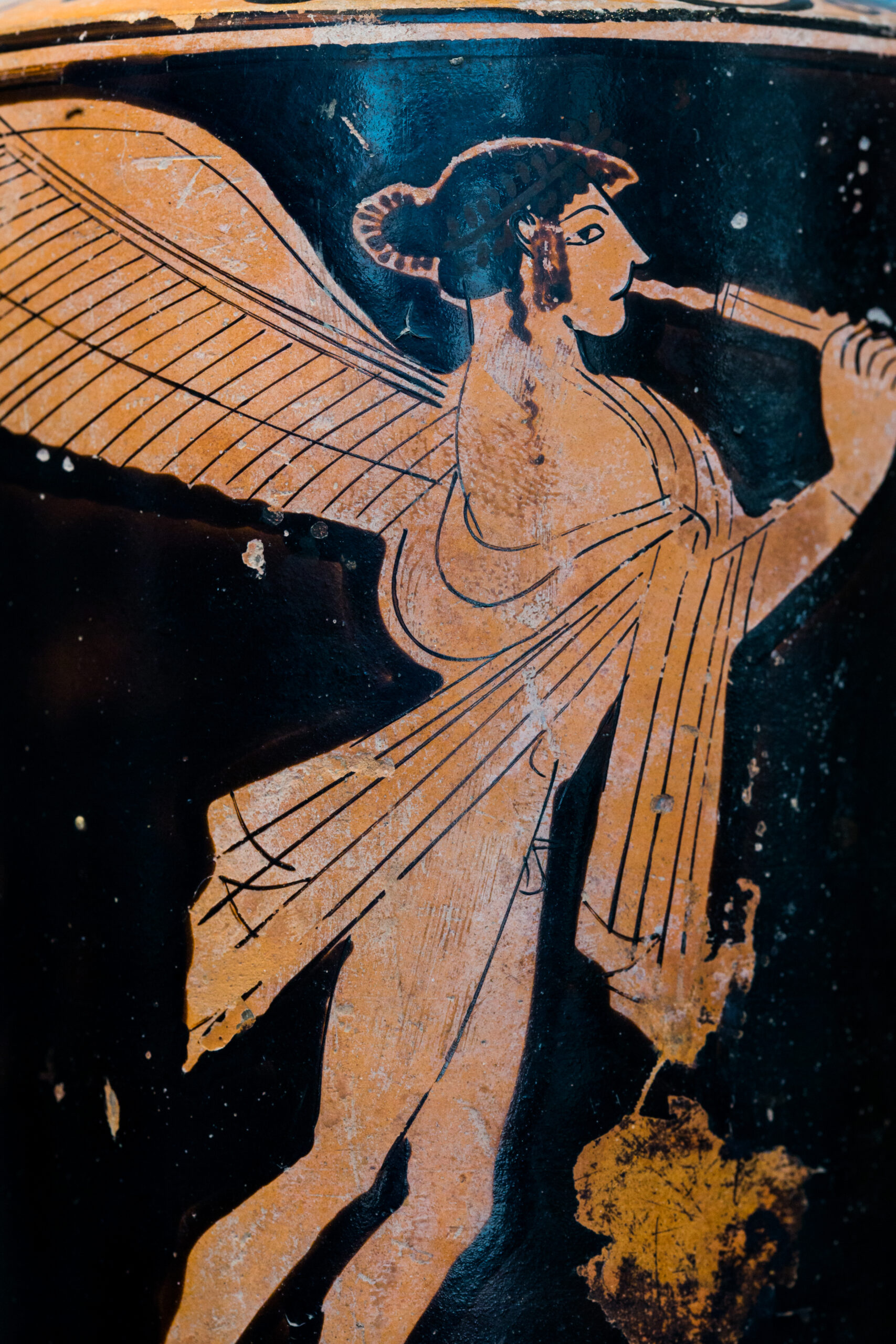 A portion of an ancient Greek red-figure vase depicting a young man with wings wearing a robe (with genitalia exposed) and playing a set of two auloi.