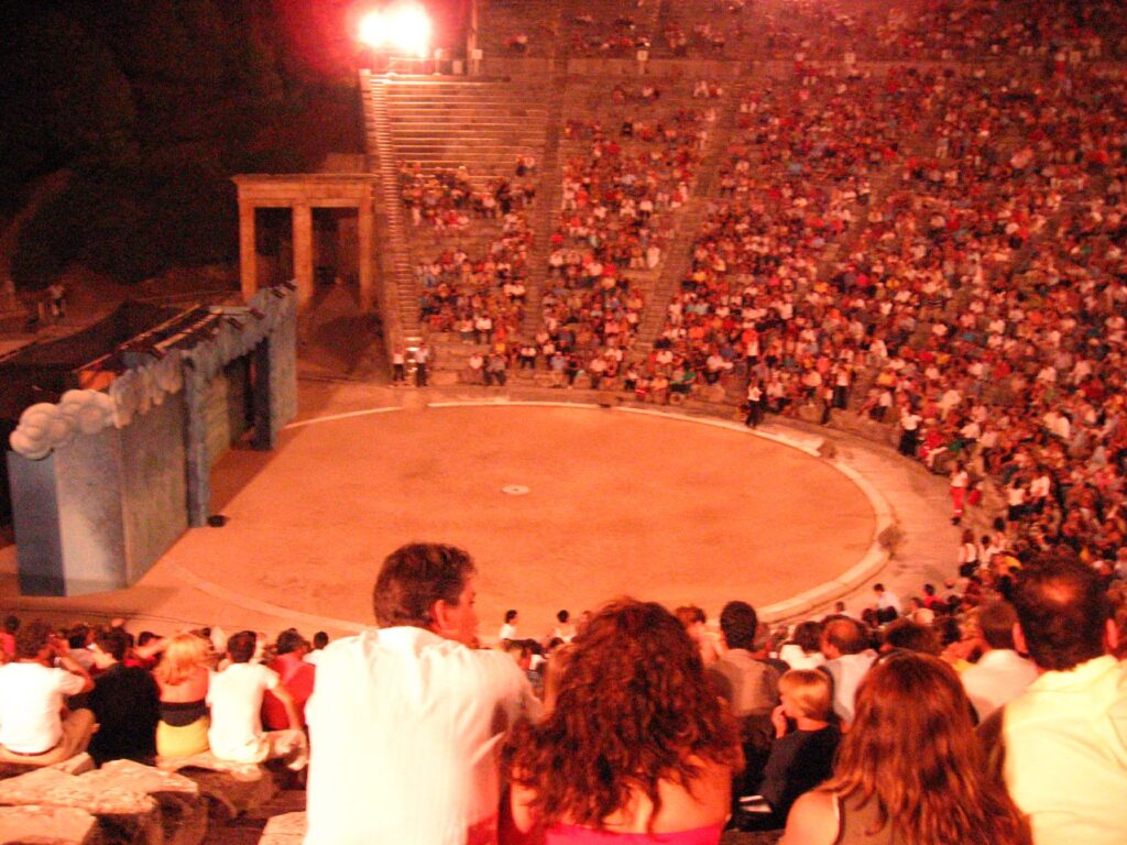 A photo of a modern audience packing an ancient theater for a theatrical production. It's night-time, and we're looking from the back of one wing of the seats, down at the circular orchestra, and with lots of seats (filled with people) arcing around it.