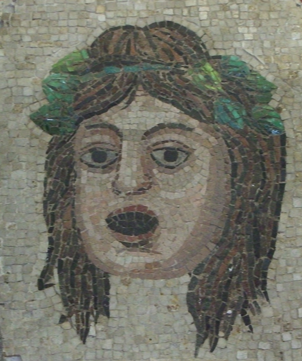 An ancient mosaic of a theater mask. The mask has pale skin tone, long straight brown hair, and a green wreath around the top of its head.