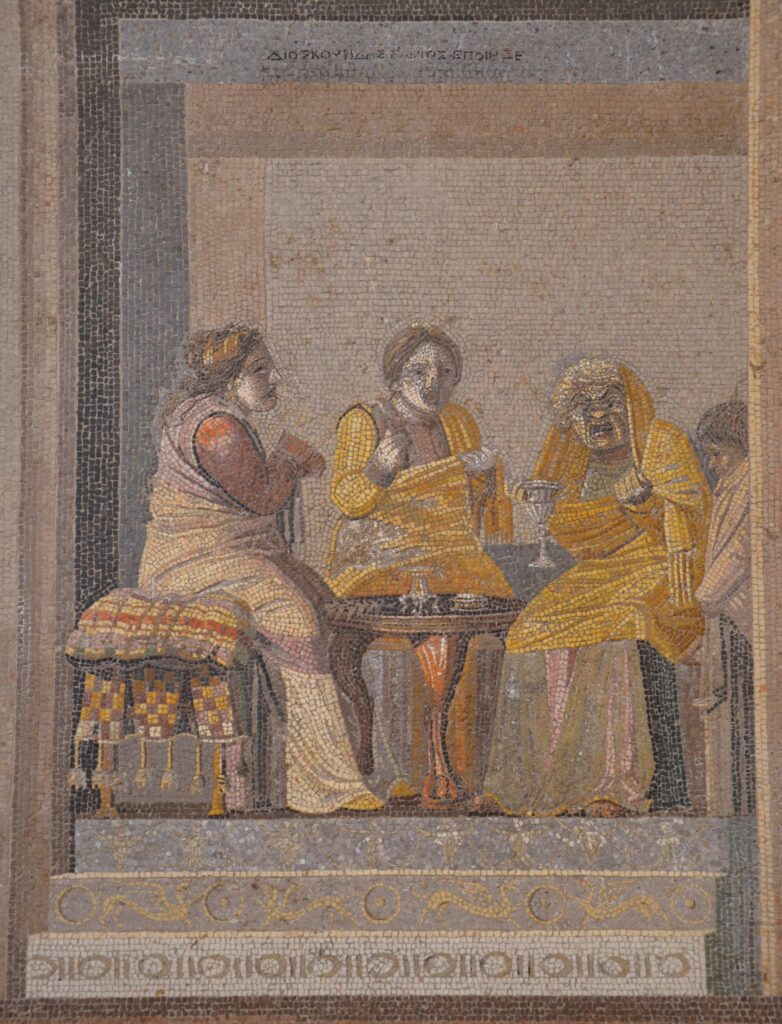 An ancient mosaic depicting a scene from a comedy. Four actors are seated on cushioned stools around a circular three-legged table. The actors are wearing full-face masks, all pale colored, two of younger women, one of an old woman, one indistinct. Two are wearing yellow robes, two beige.