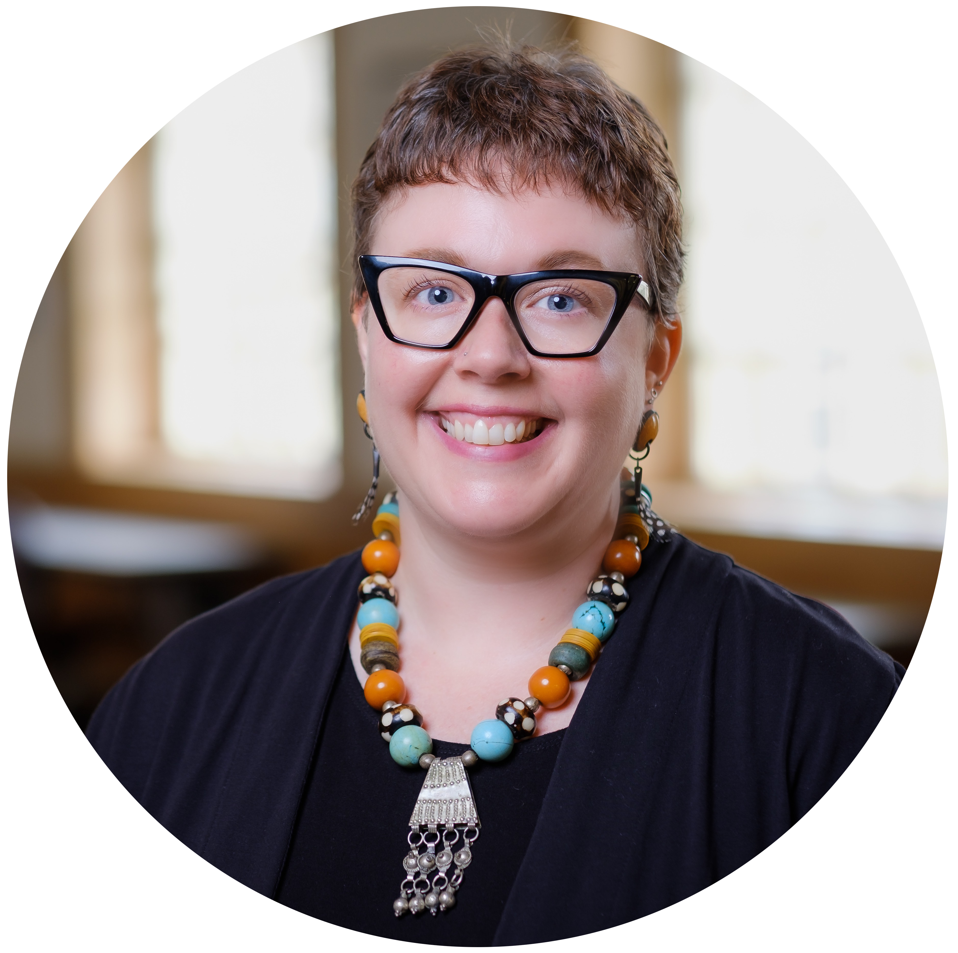 Profile picture of Molly Keener: a white woman with short-cropped brown hair and glasses. She is smiling at the camera. She is wearing a black shirt, black jacket, and big necklace with large bright blue and orange beads and a dangly silver centerpiece.