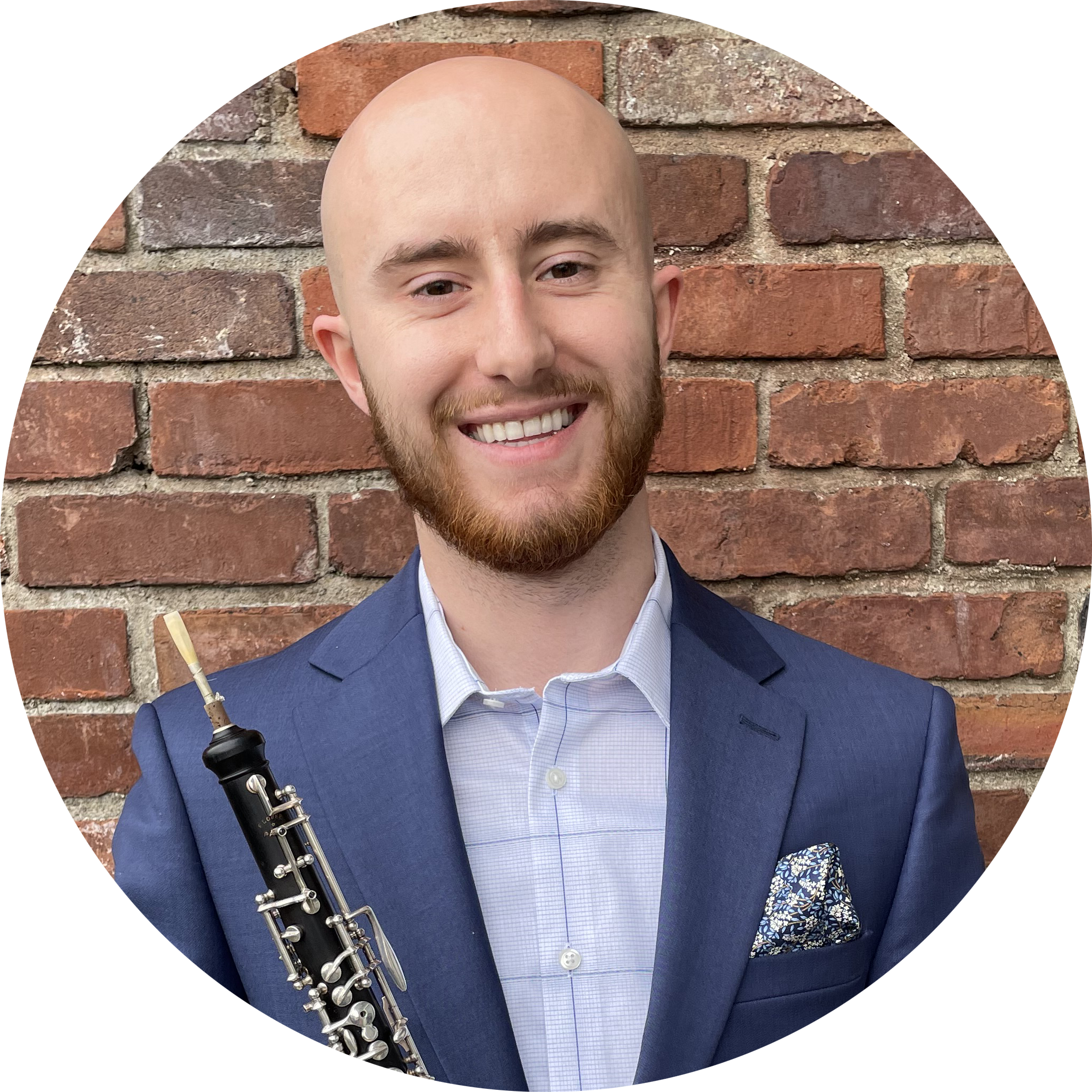 Profile picture of Dane Bennett: a white man with clean-shaven head and a brown beard. He is smiling and wearing a light blue button-up shirt, a darker blue sport coat, and a blue-and-white pocket square. He is holding an oboe.