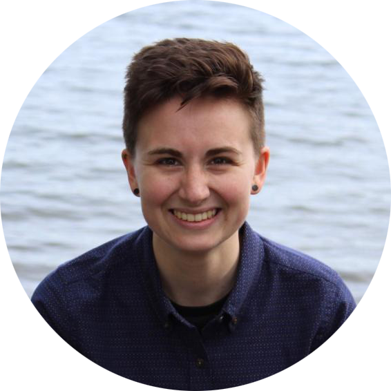 Profile picture of Sarah Brucia Breitenfeld: a white person with short brown hair. They are smiling and wearing a dark blue button-up shirt. Behind them is open water on a calm day.