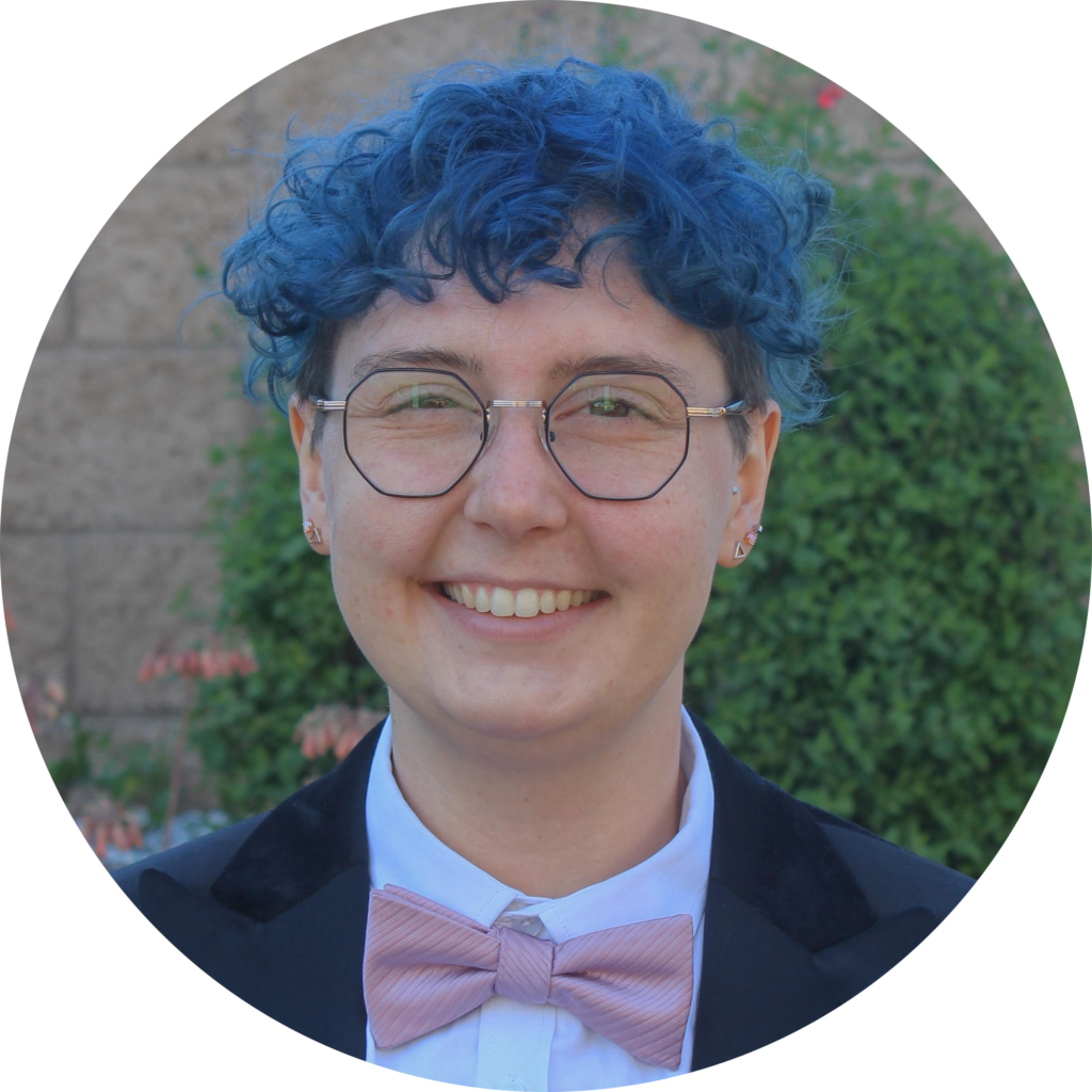Profile picture of Cypris Roalsvig: a white person with short bright blue curly hair and octagonal glasses. They are smiling and wearing a white button-up shirt, navy blazer, and pink bowtie.