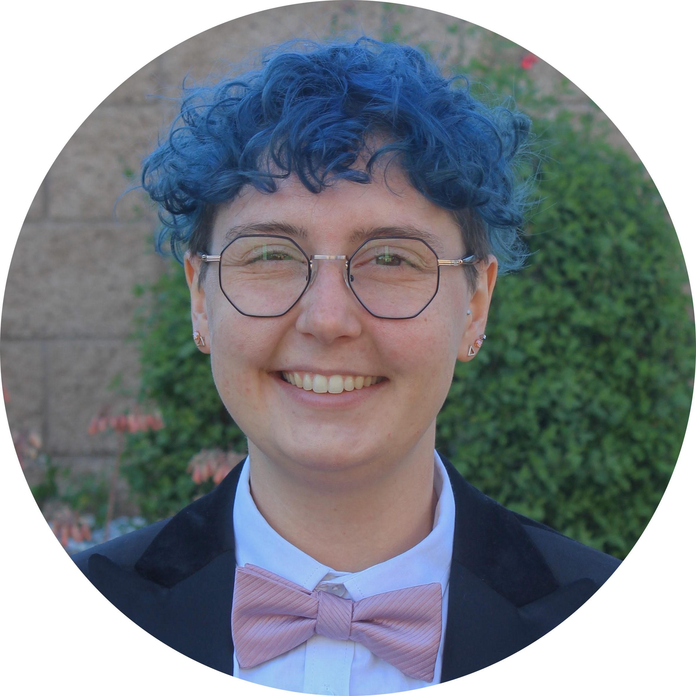 Profile picture of Cypris Roalsvig: a white person with short bright blue curly hair and octagonal glasses. They are smiling and wearing a white button-up shirt, navy blazer, and pink bowtie.