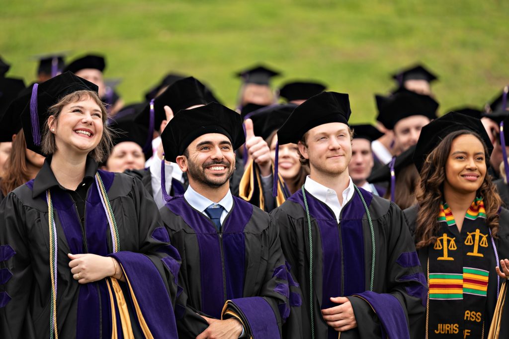 four graduates, a young woman, then two young men, then another young woman look up into the distance. They are wearing their graduation robes and caps. The young woman on the right is wearing a Black Law Students Association sash. They are smiling.