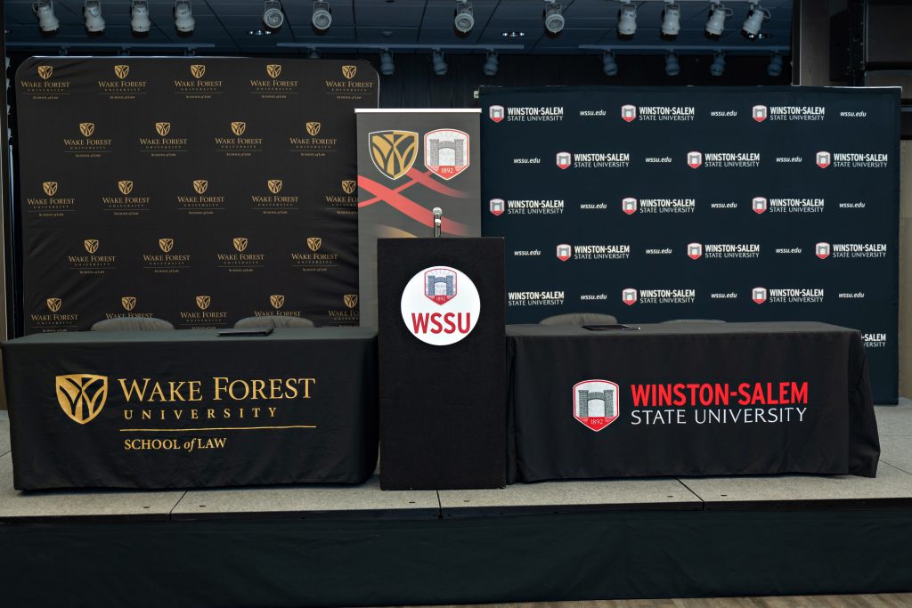 The stage decorated with Wake Forest Law and WSSU backdrops, two tables, one with a Wake Forest Law tablecloth and one with a WSSU tablecloth, as well as a vertical banner that combined both universities logos, and a WSSU-branded podium