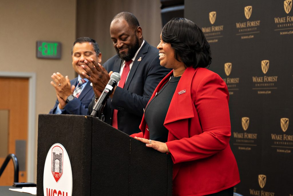 WSSU Chancellor Bonita Brown smiles at the podium as WSSU Provost Anthony Graham and Wake Forest Chief Diversity Office Jose Villalba clap