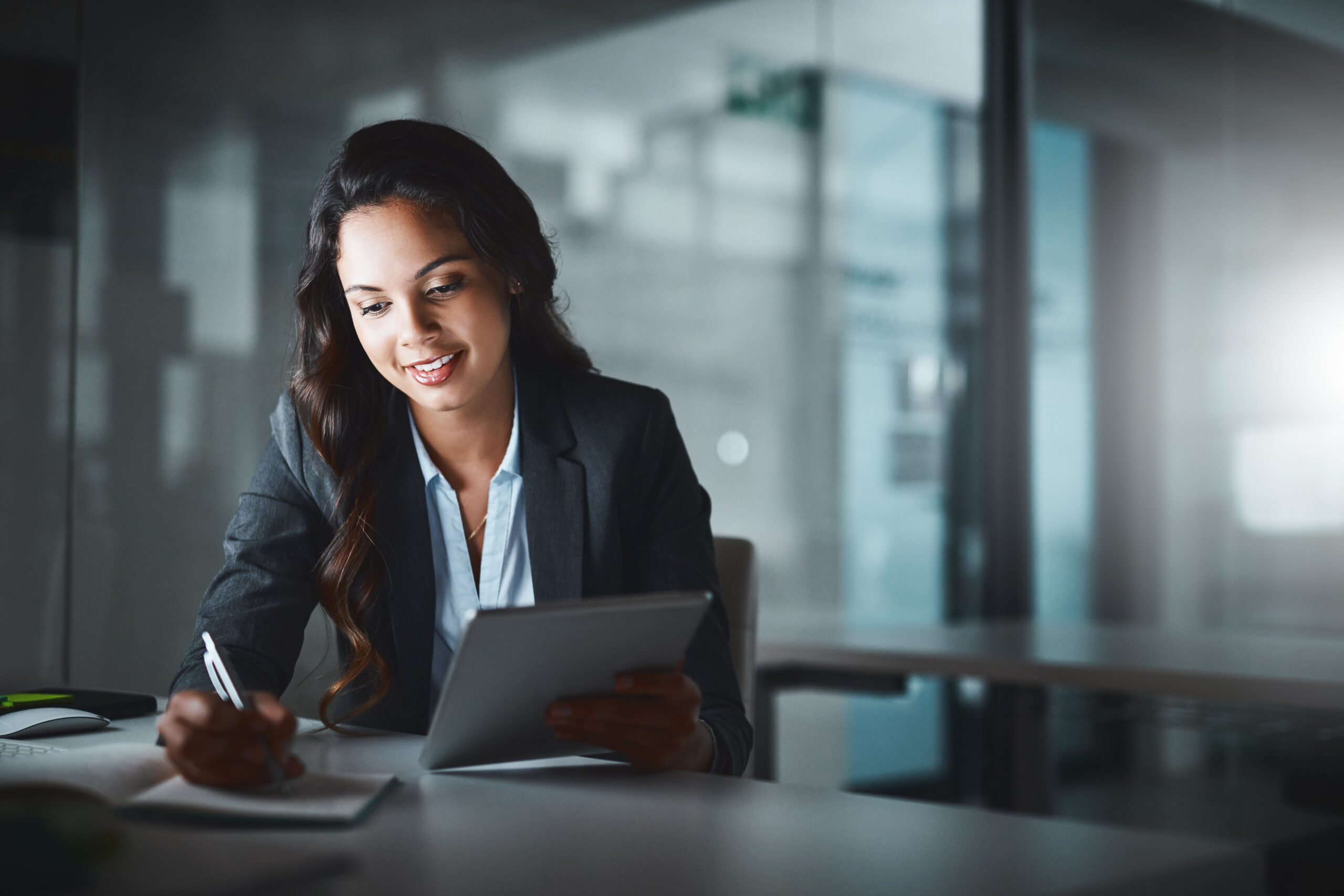 business woman sitting at desk writing in a notebook and holding a tablet