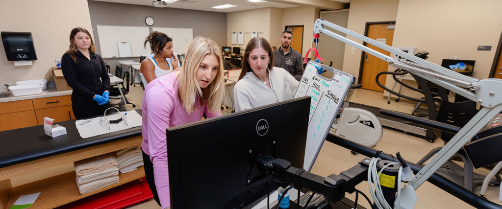 Students at computer in lab, with faculty and other students in background
