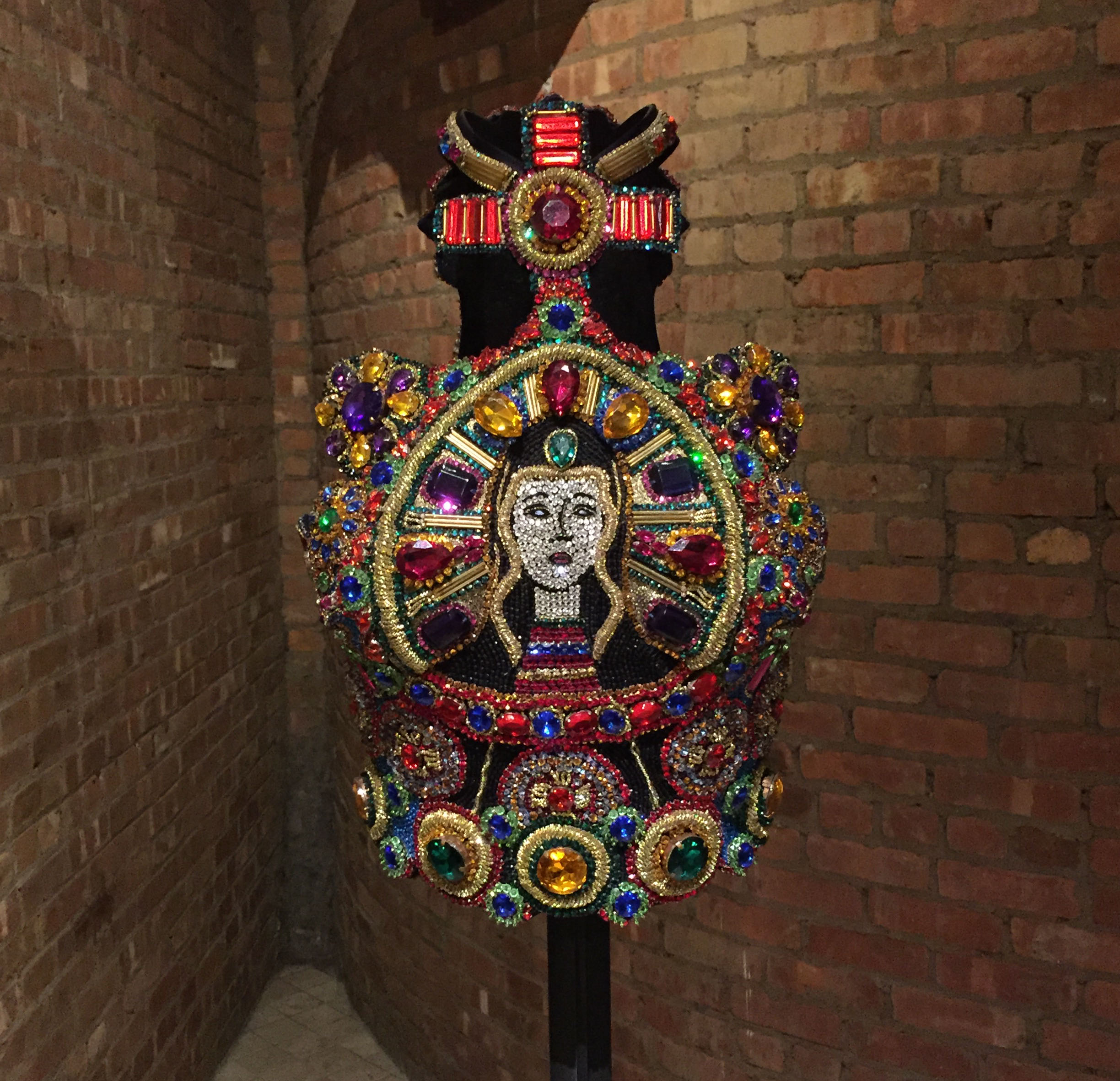 The shape of the halo, anchored by a beaded band at the bottom, resembles that of a mitre, a ritual cap worn by Catholic religious leaders (bishops, cardinals, the Pope). Atop this mitre shape, the beaded neckpiece forms the shape of a cross made from a mosaic of red gems. The overall effect is stunning and exuberant--an explosion of color and a celebration of Mary’s status in Catholicism and in fashion.