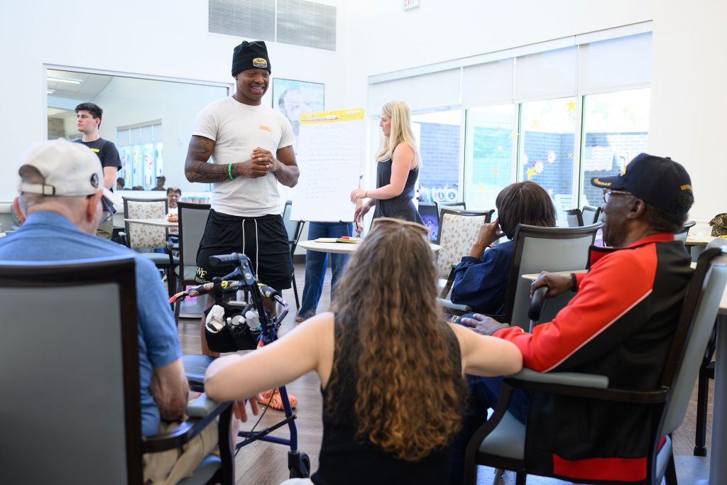 A student stands in front of a group of elderly in wheelchairs as he presents his findings.