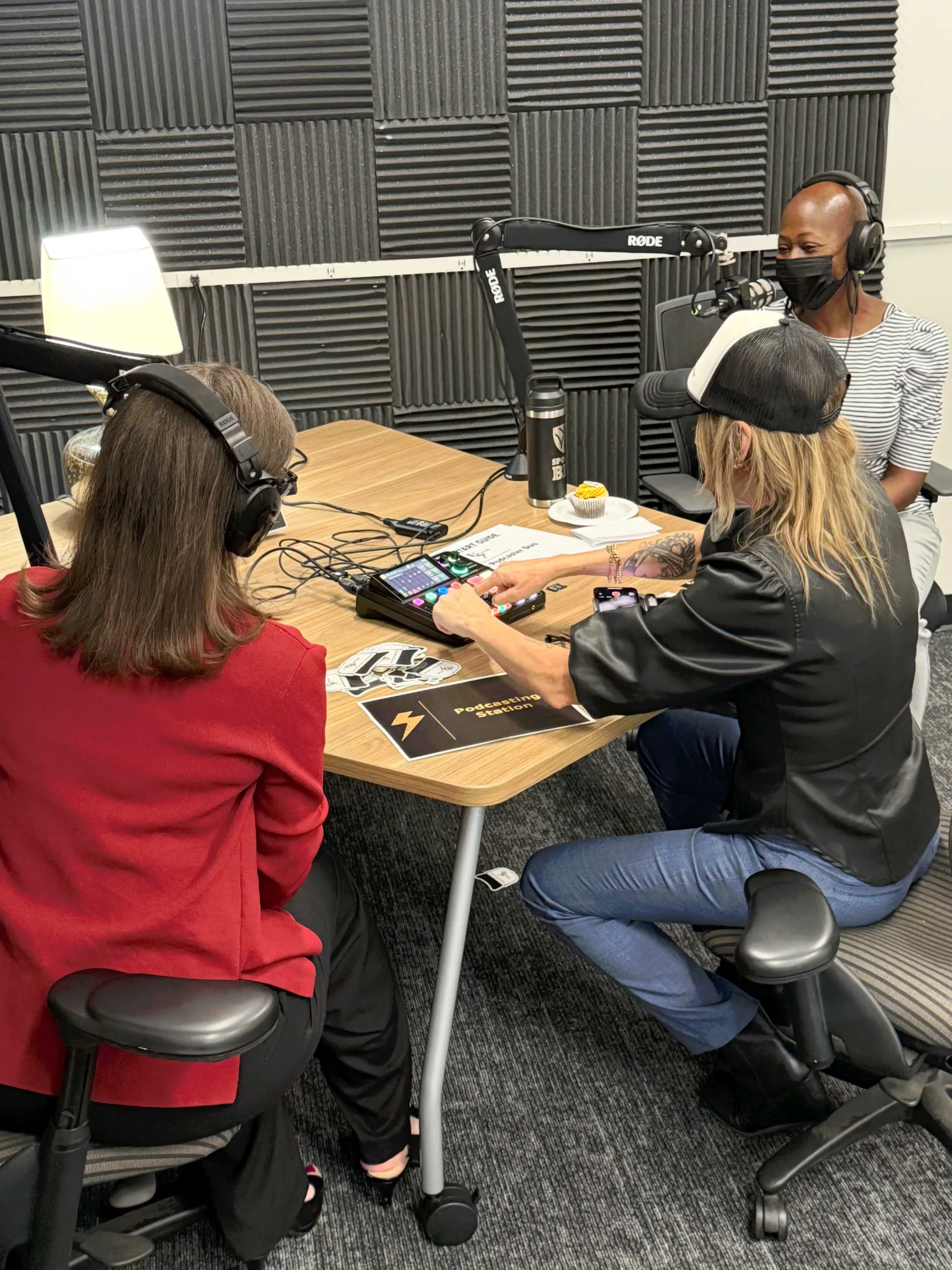 Brianna Healey runs the mixer at the podcasting station while a faculty member in a red blazer sits across the table wearing headphones.