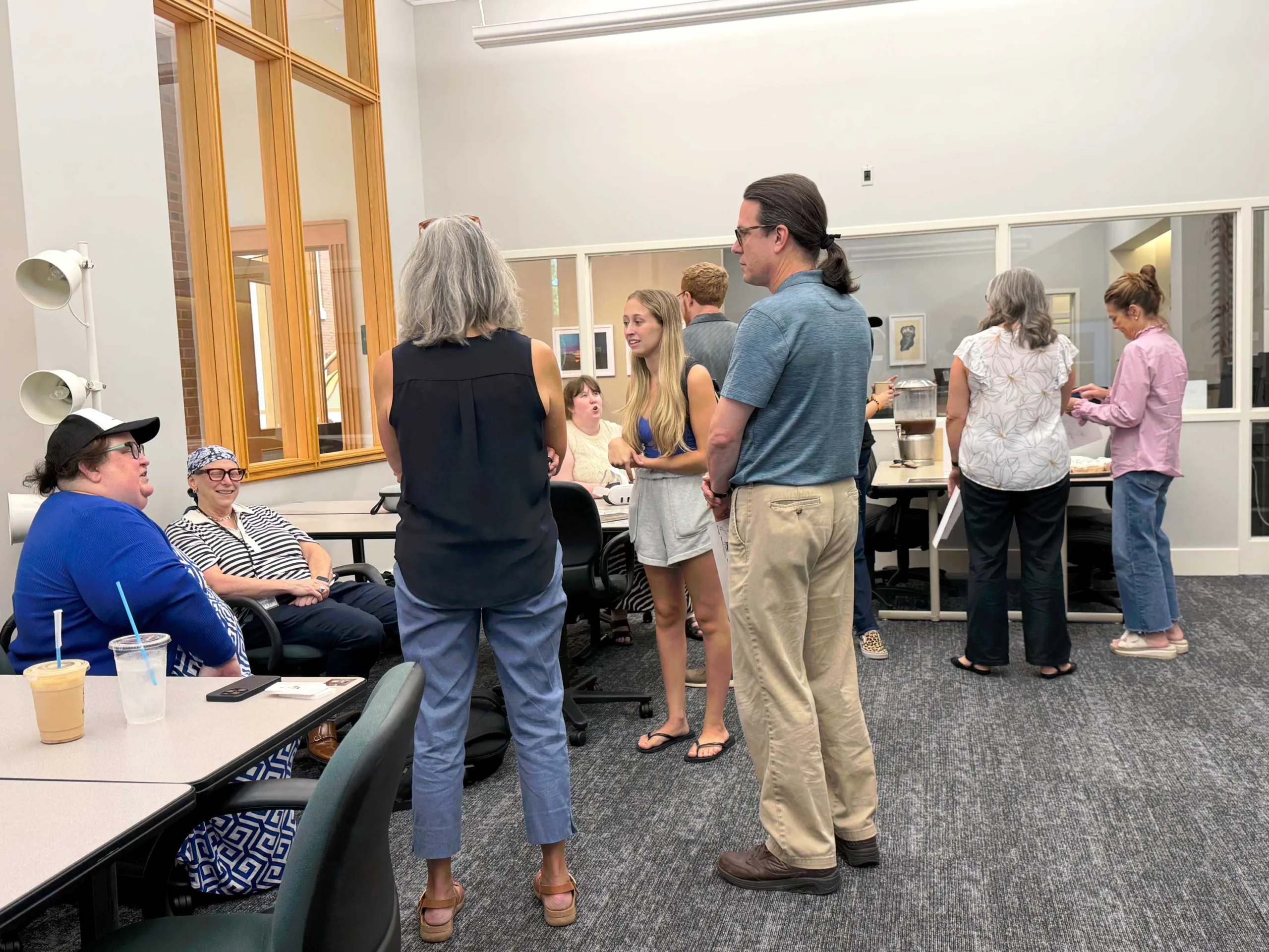 Guests stand and sit in conversation in a library meeting room with glass walls, chairs, and a refreshment table in the background.