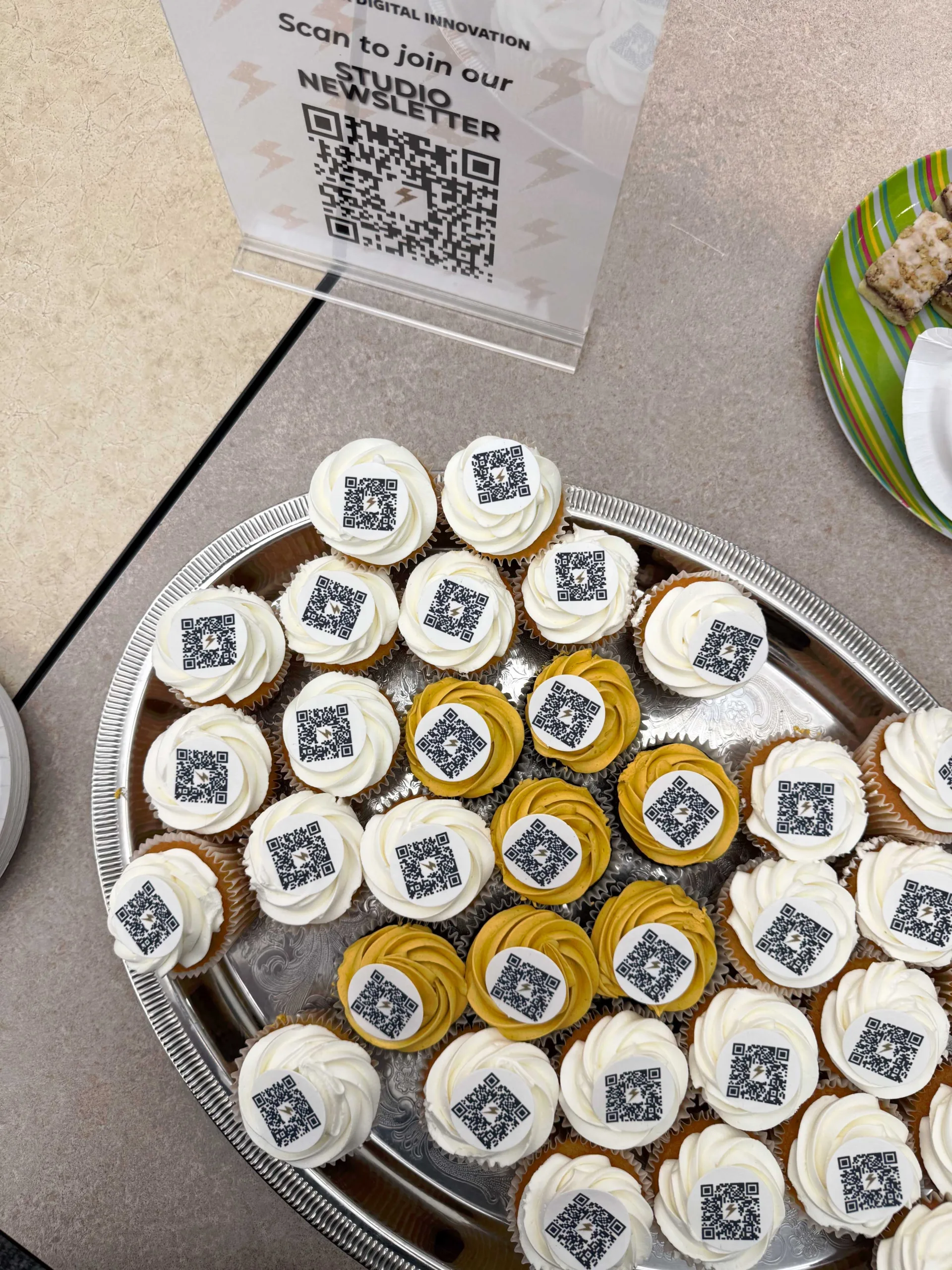 A tray of cupcakes, each topped with a QR code, sits beside a sign reading "Scan to join our Studio Newsletter."