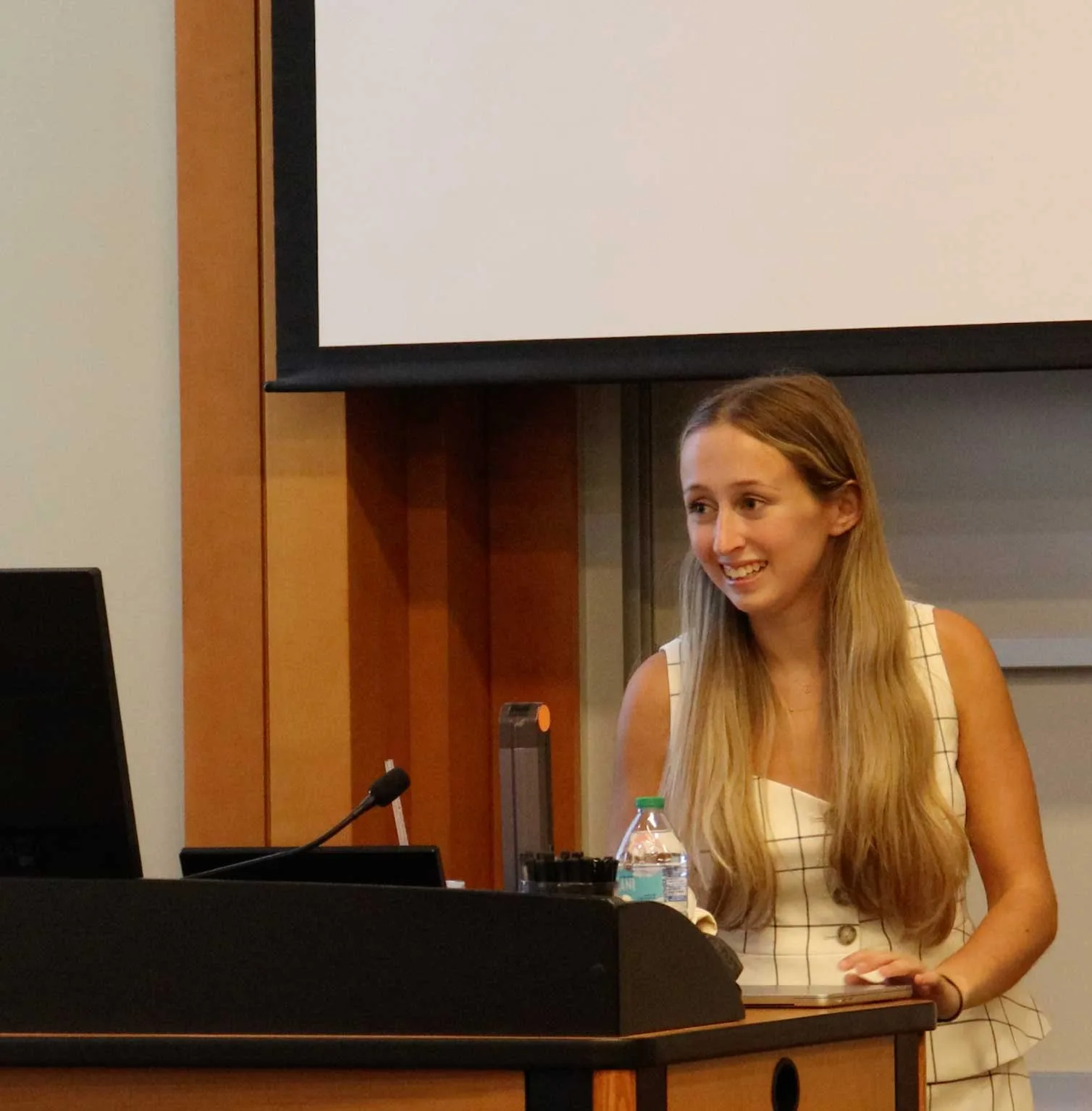 Lyla Holstein smiles as she sits at the front of the room with a microphone and laptop in front of her and a projection screen behind her.