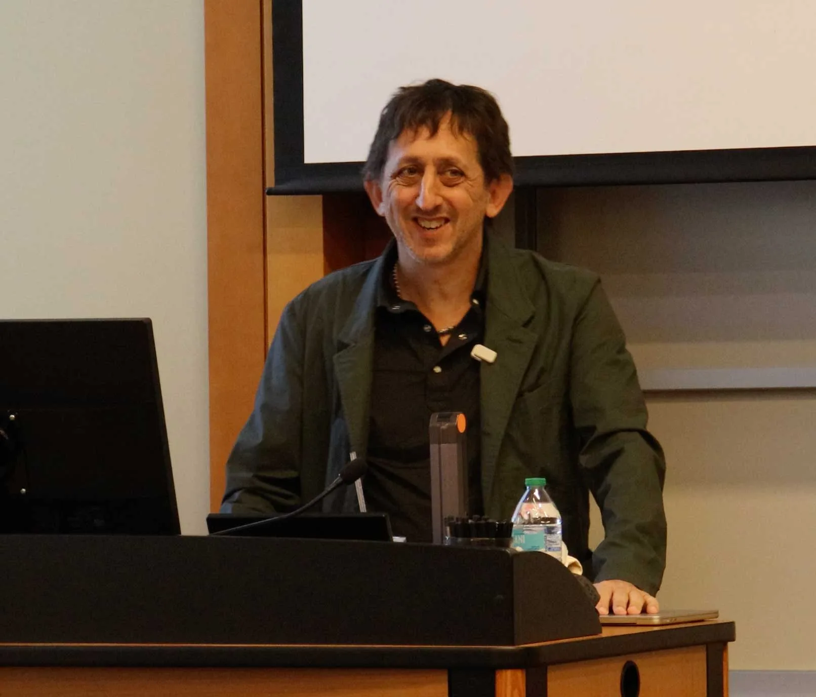 Michael Holstein smiles as he speaks from a podium with a microphone and laptop, with a projection screen behind him.