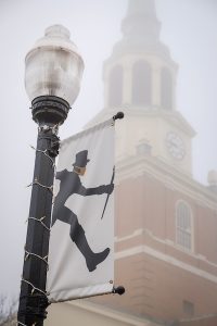 A light pole banner showing the profile of the Deacon wearing a mask. Behind the banner, Wait Chapel is seen through the fog