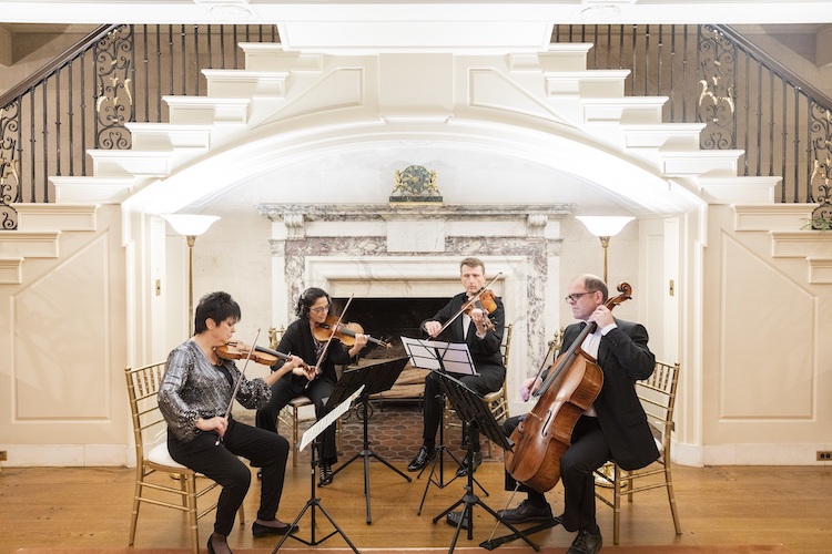 Photo of three musicians seated playing violins and one seated playing cello in front of a grand double staircase