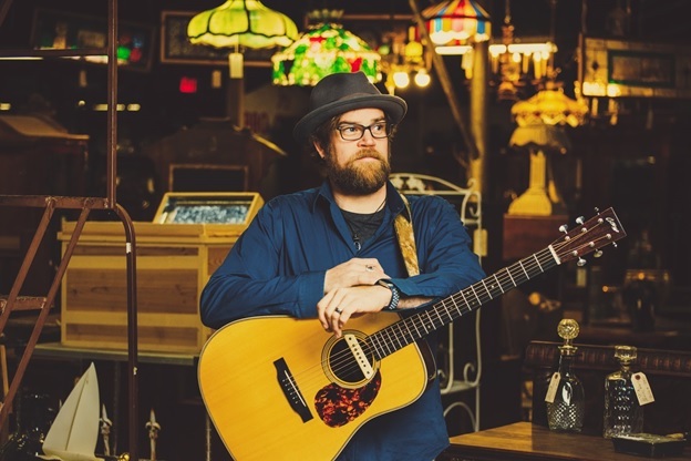 Photo of musician Aaron Burdett with his guitar standing an a cozy music hall